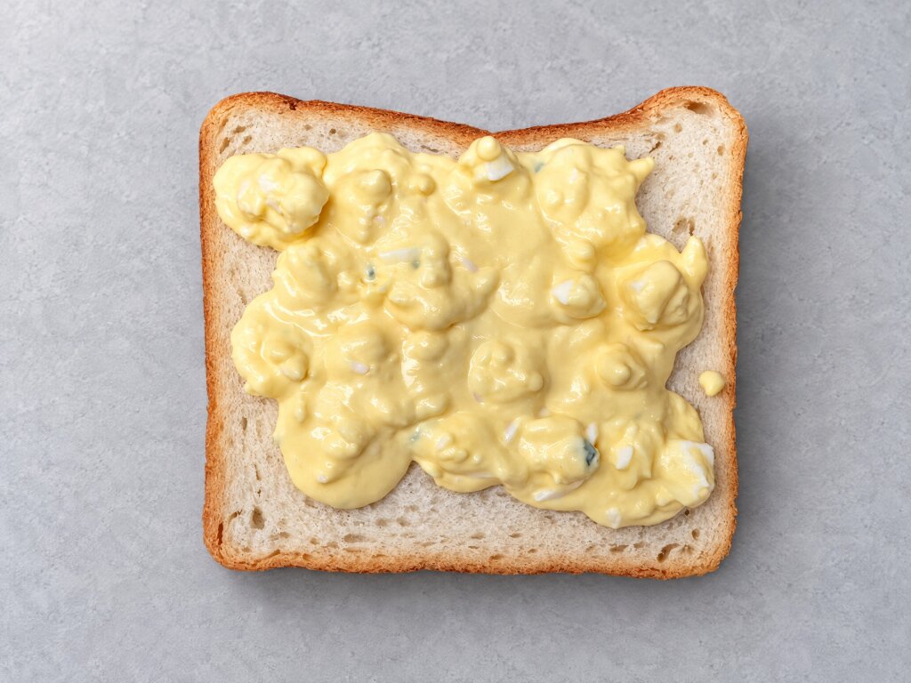 Slice of white bread with a layer of egg salad deposited on it, top view