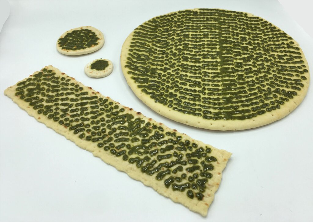 Green pesto deposited in short lines on a large round base, a long flatbread and two small round flatbreads, on a white surface