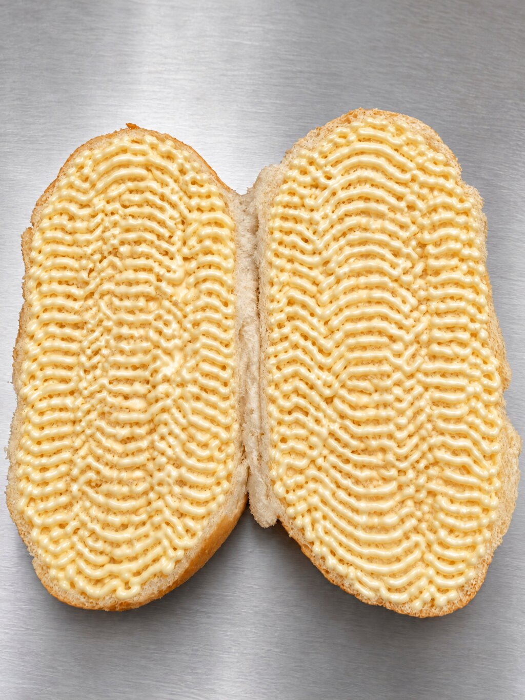 Two halves of a bread roll with wavy lines of mayonnaise across the whole cut surface, up to the crust
